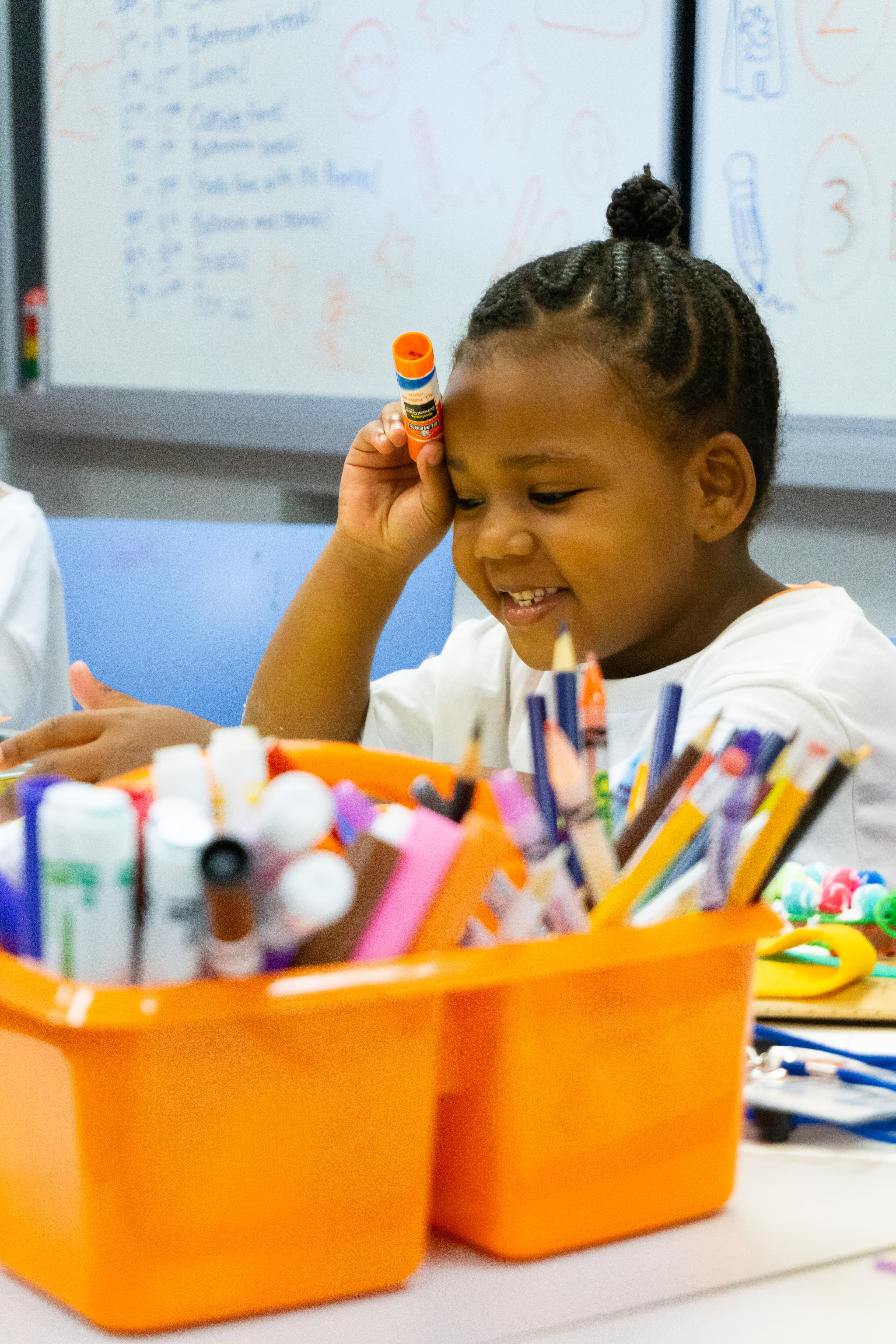 Child sitting holding glue stick in front of art caddy smiling.