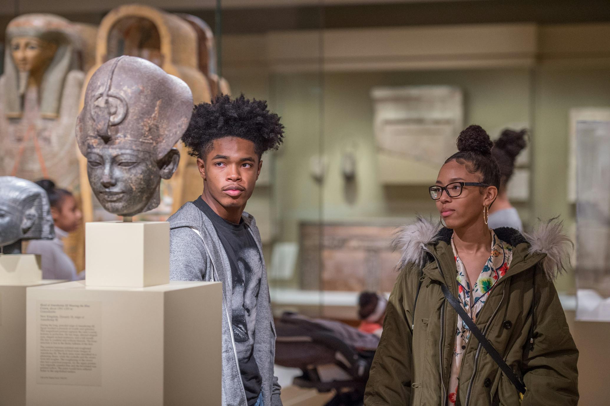 two visitors look at dark stone bust of figure with egyptian headdress on pedestal in gallery