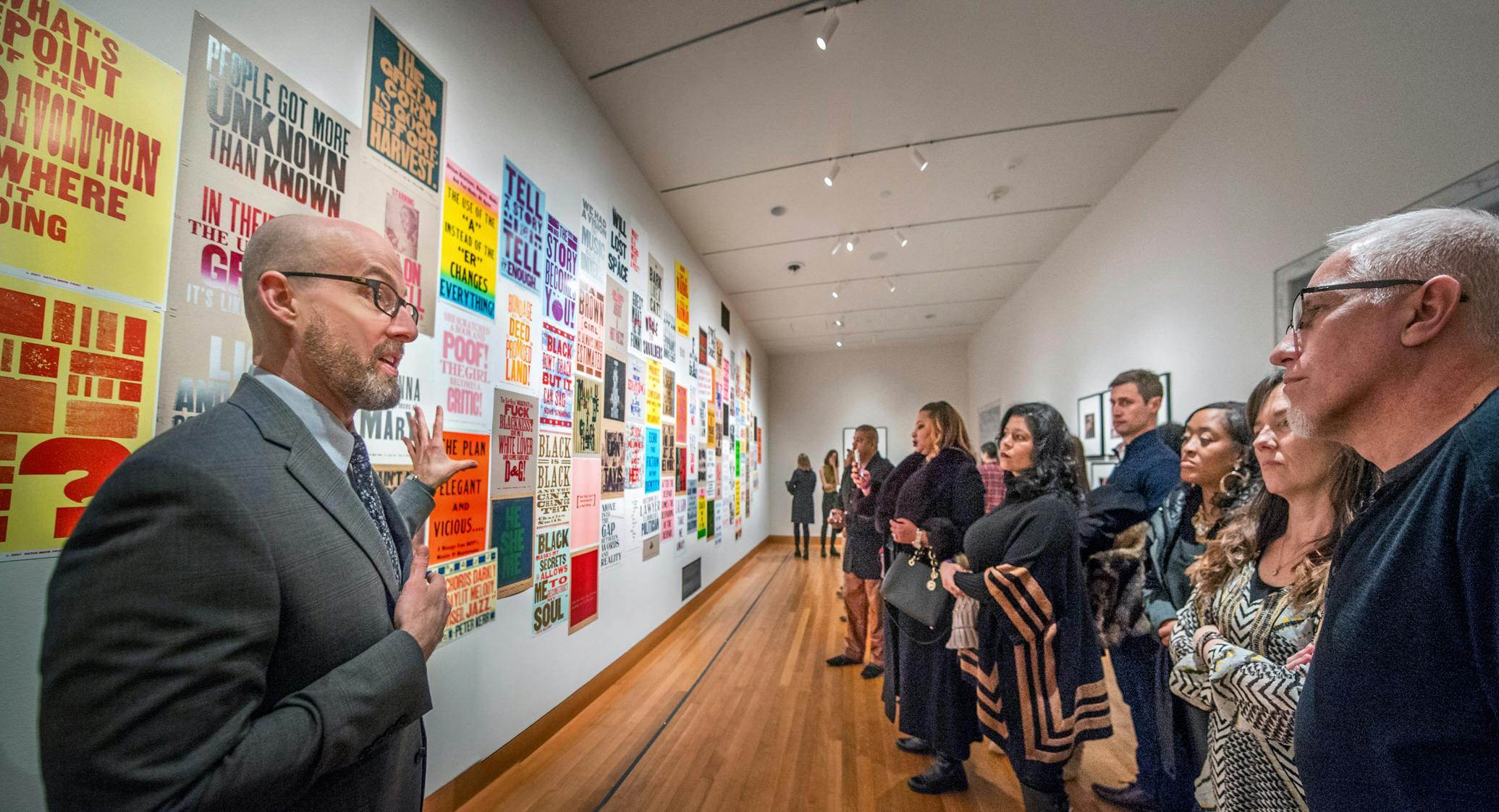People in gallery listen to tour guide in front of wall covered in brightly colored graphic posters