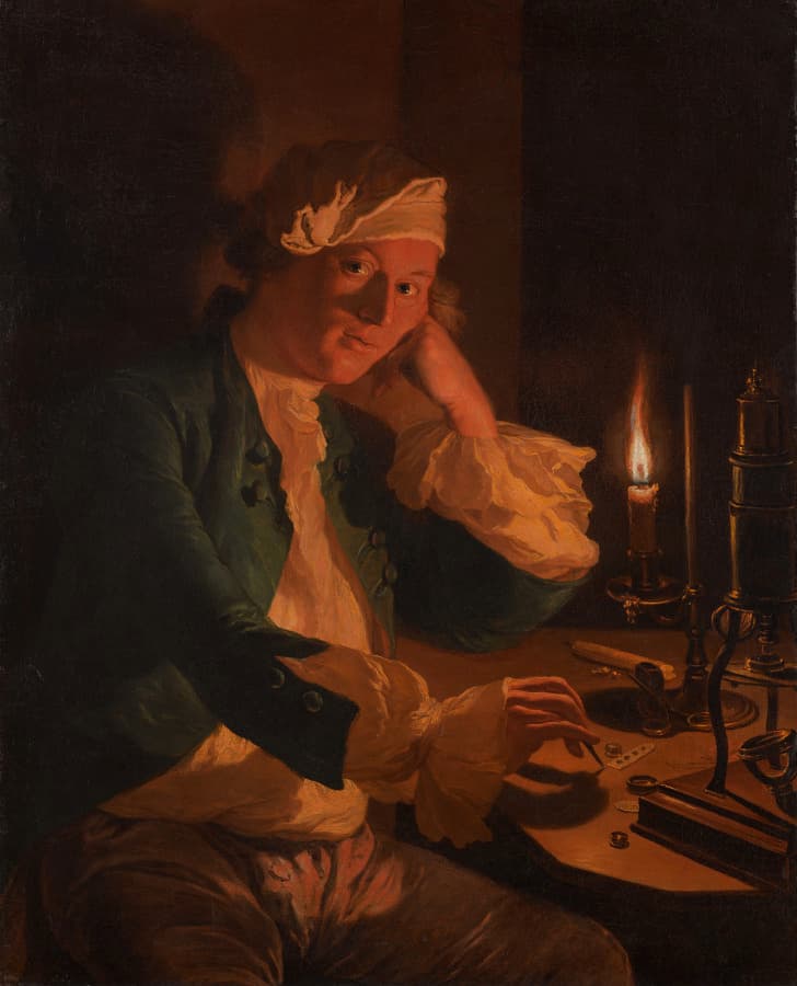 A man lit by a single candle wearing a cap sits at desk, His face rests on his hand, the direction of his gaze appears to be aimed back at the viewer.  